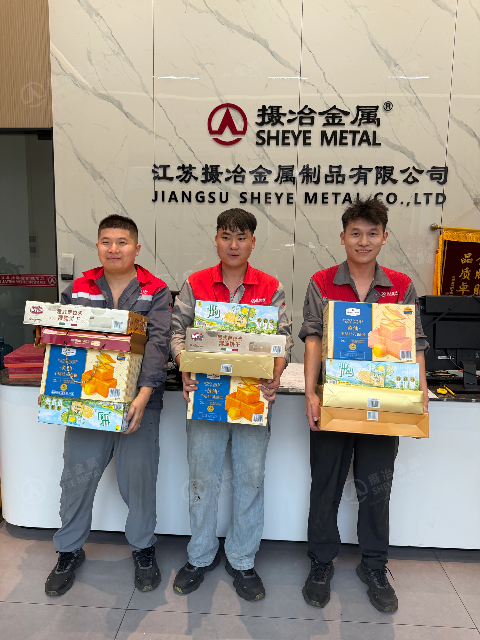 Three Sheye Metal colleagues in red and grey workwear holding gifts beneath the company sign.